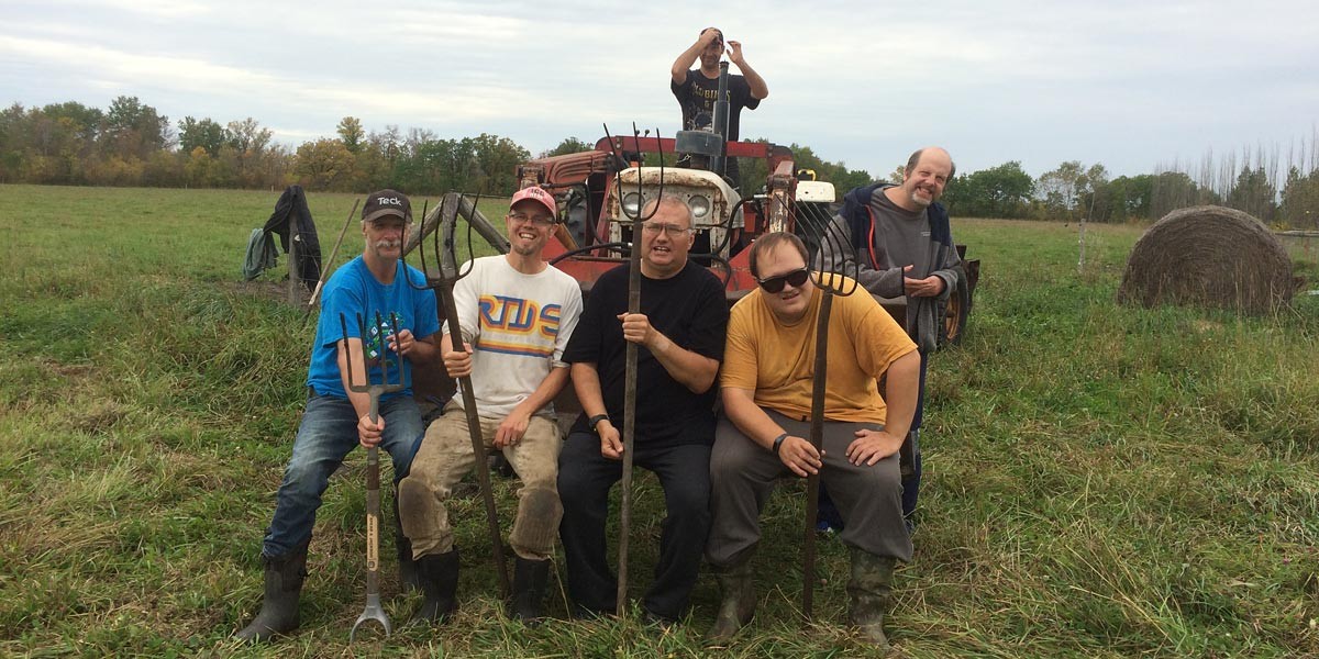 Six men posing with pitchforks and tractor in a grassy field with hay bale and trees in background.