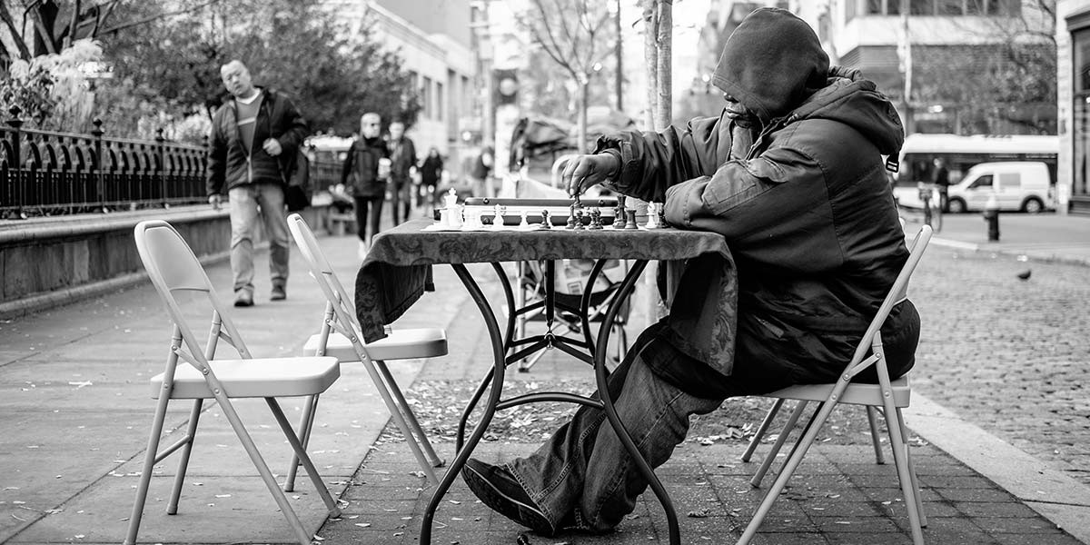 Person in hooded jacket playing chess alone at a table on a city sidewalk with empty chairs.
