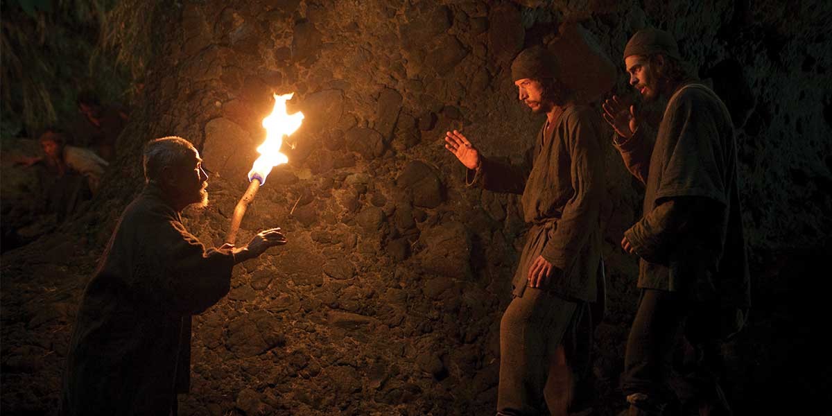 Three men in dark clothing around a lit torch against a rocky wall in a dim setting.