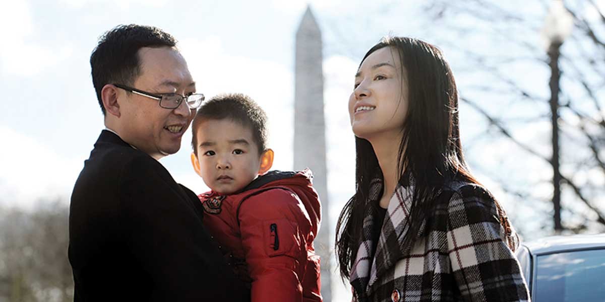 Family of three outdoors with Washington Monument blurred in the background on a sunny day.