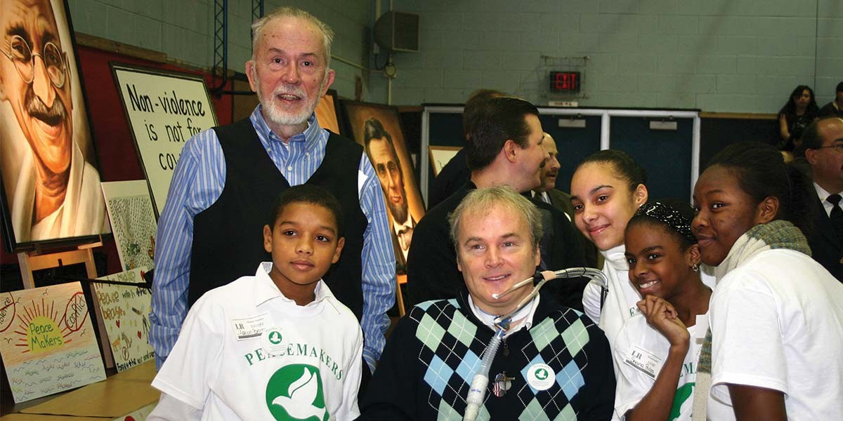 Group photo of diverse children and adults with peace-themed shirts and posters in a gymnasium.