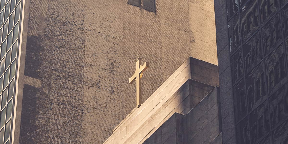 Golden cross atop a concrete building with brick and glass buildings in the background.