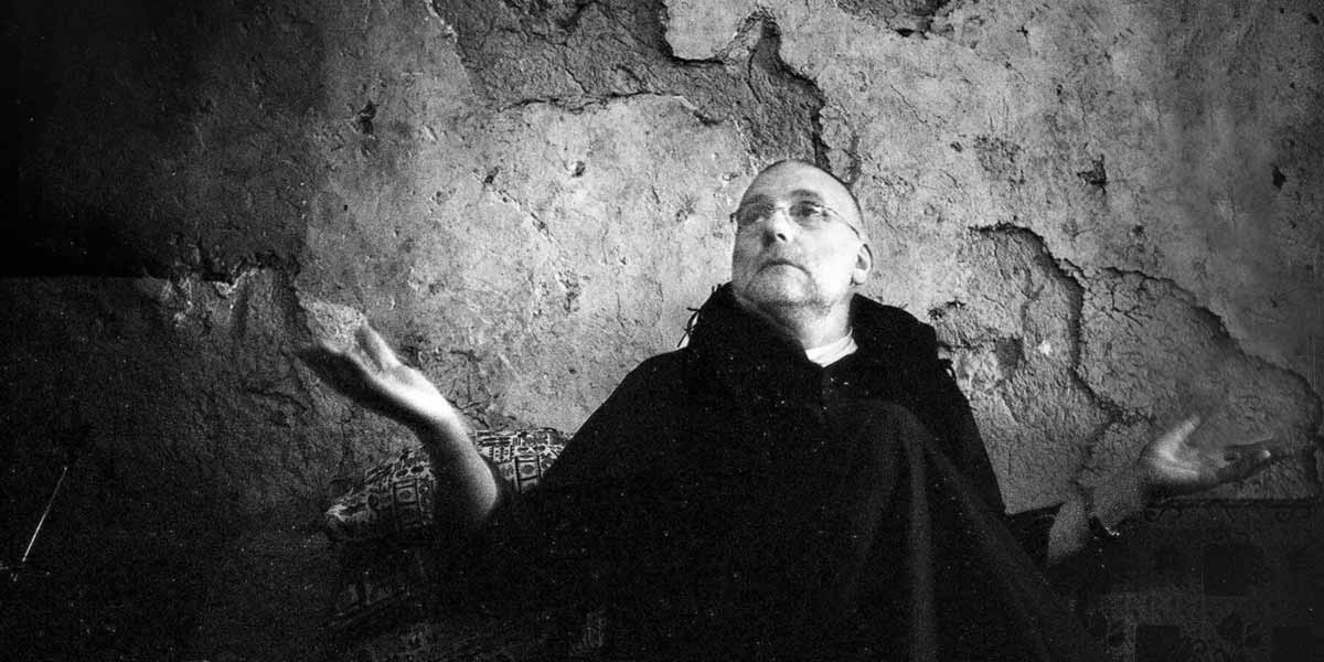 Black and white photo of a person in glasses raising hands against a textured, cracked wall.