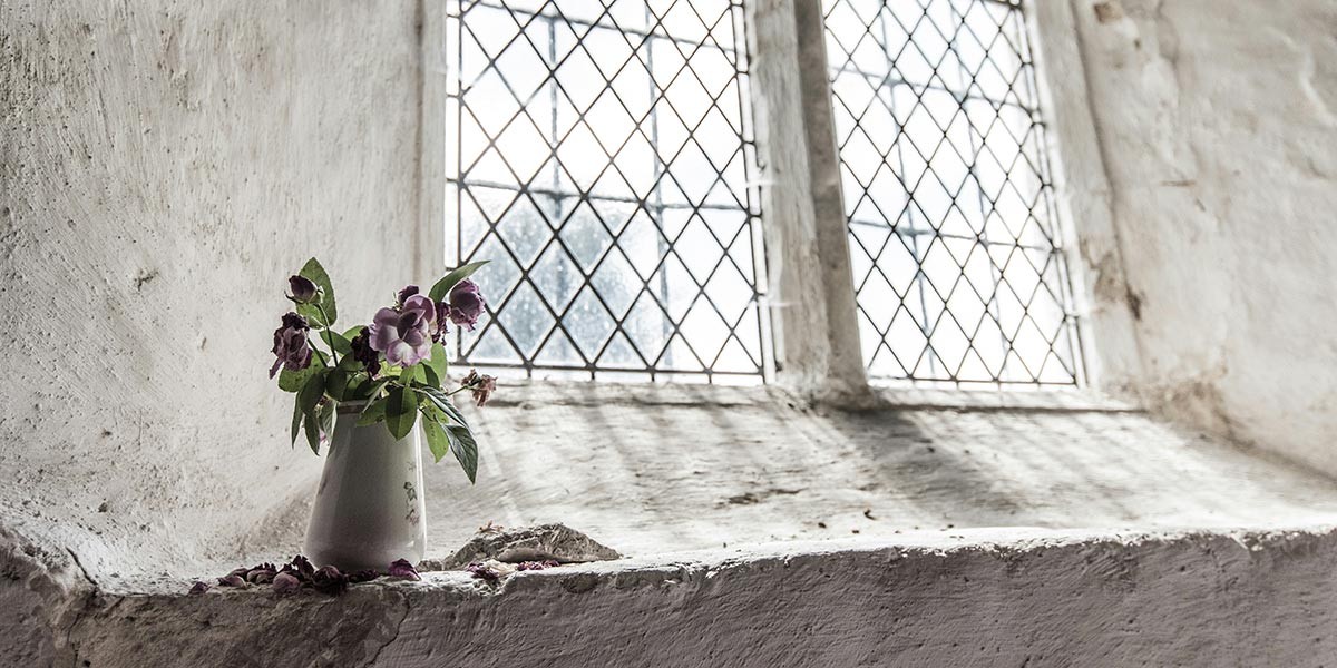 White vase with purple flowers on a rough stone windowsill with leaded glass windows.