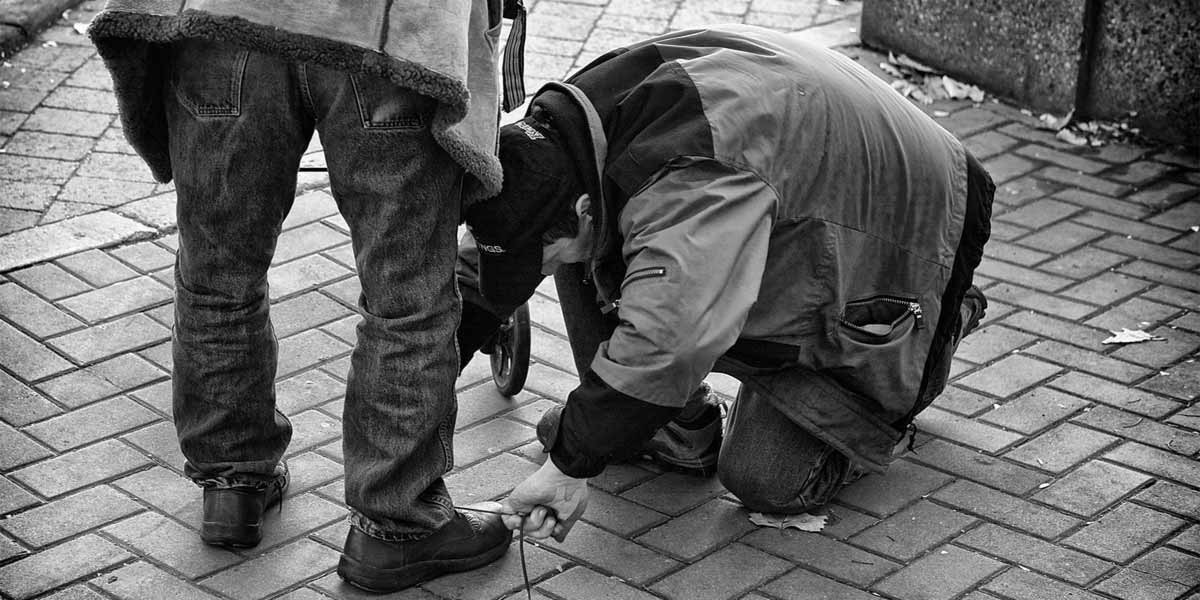 Person kneeling on sidewalk tying another person's shoelace on a brick paved pavement.