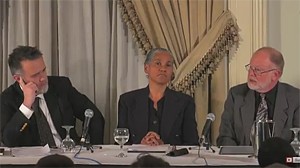 Three men in suits seated at a panel table with microphones and water glasses during a discussion.