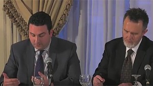 Two men in suits seated at a table speaking into microphones during a formal event.