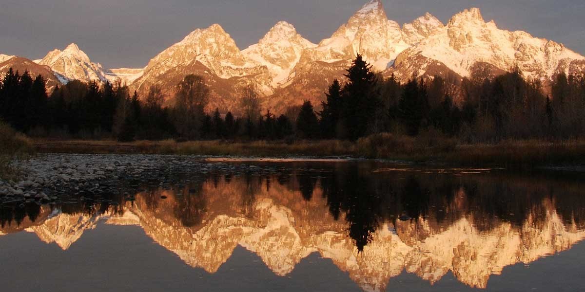 Snow-capped mountain range reflected in a calm river with dark pine trees in the foreground.