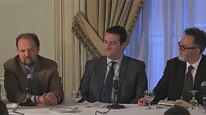 Three men in suits seated and engaged in discussion at a panel table with microphones and glasses.