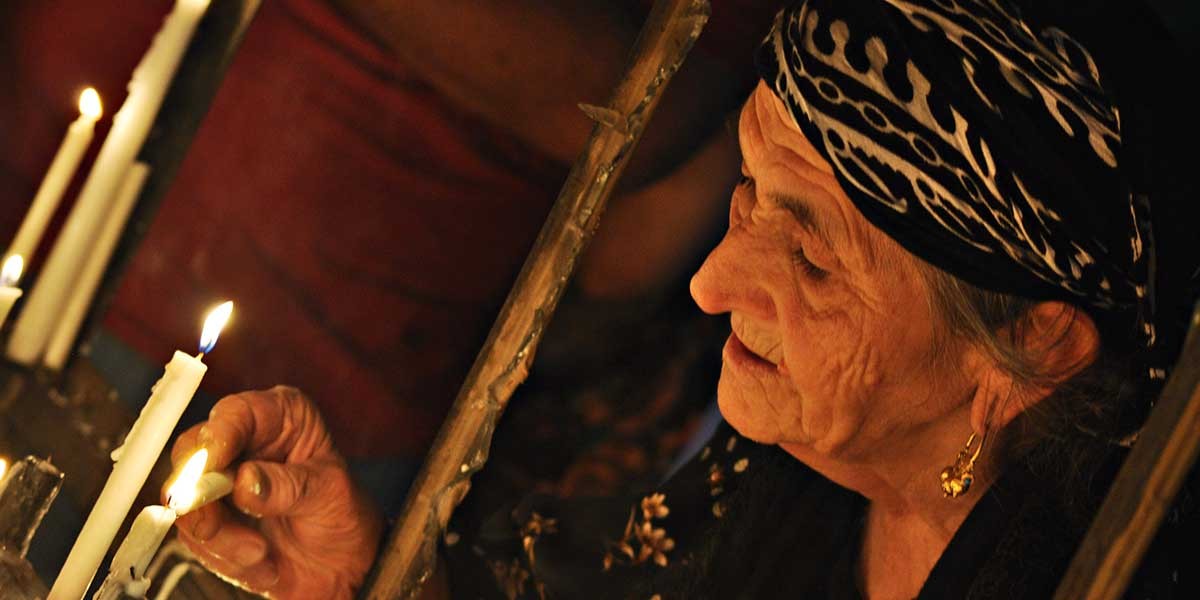 Elderly woman lighting candles wearing a black headscarf with white patterns and gold earrings.
