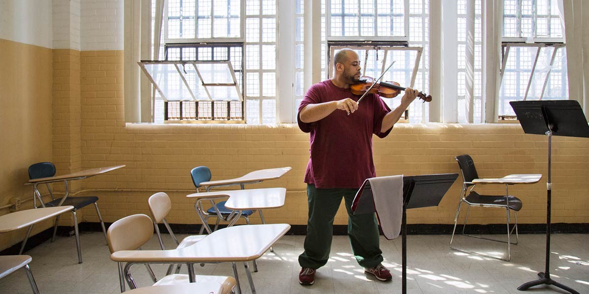 Man playing violin in a classroom with desks and open windows in the background.