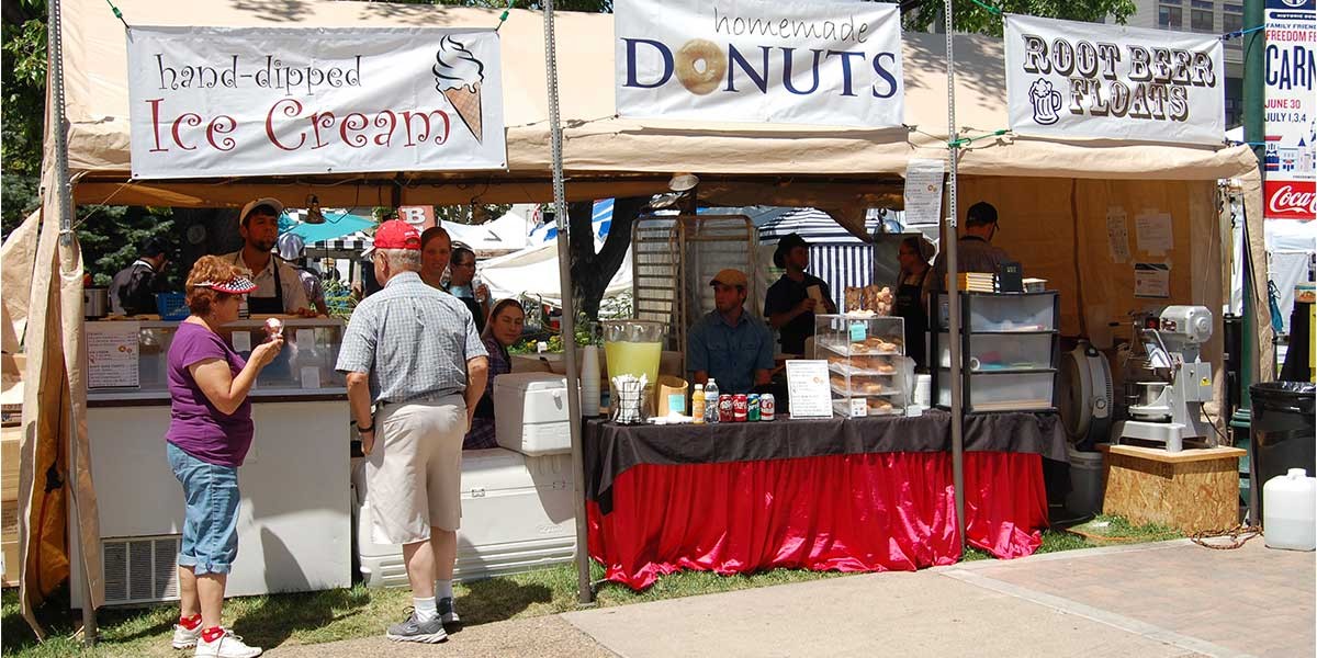 Outdoor market booth selling hand-dipped ice cream, homemade donuts, and root beer floats with customers.