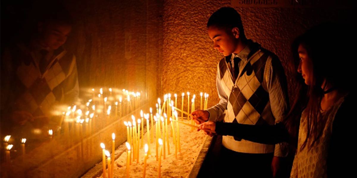 Two people lighting candles together in a dimly lit space, their reflections on nearby glass.