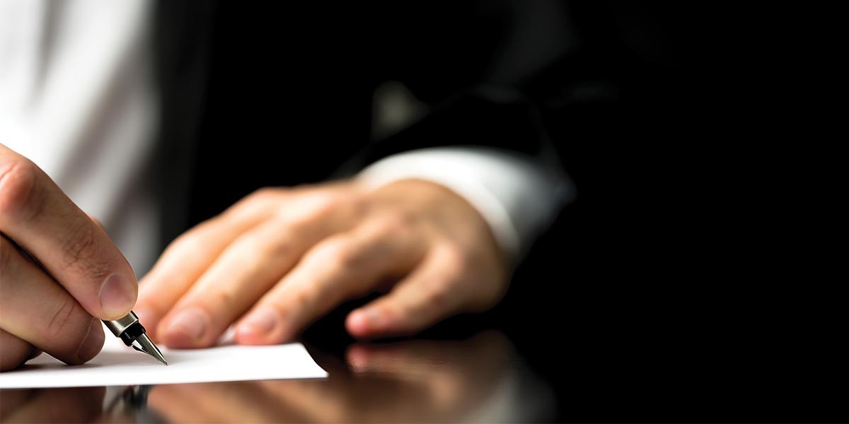 Hand holding a fountain pen poised to write on white paper on a reflective surface.