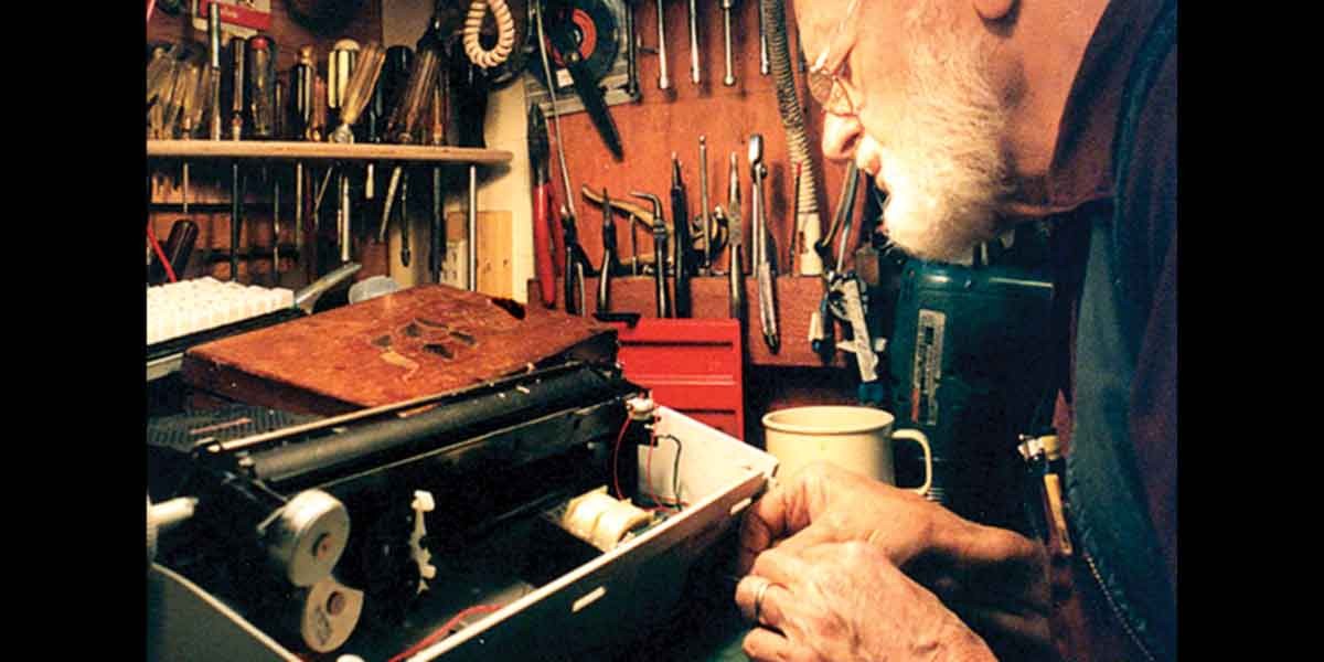 Elderly man with glasses repairing a vintage typewriter at a workbench with tools on the wall.