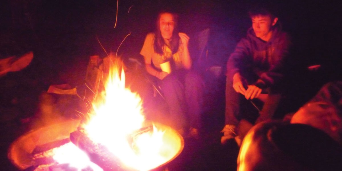 Teenagers sitting around a campfire at night, with bright flames lighting their faces.