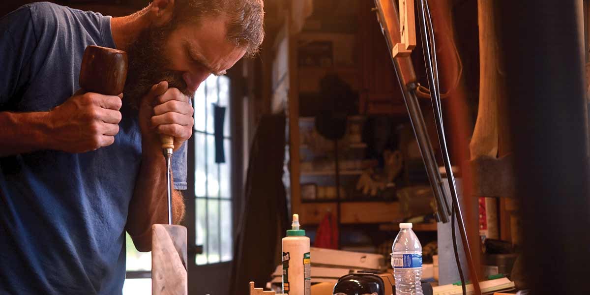 Man with a beard carving wood with a chisel in a workshop with tools and bottles nearby.