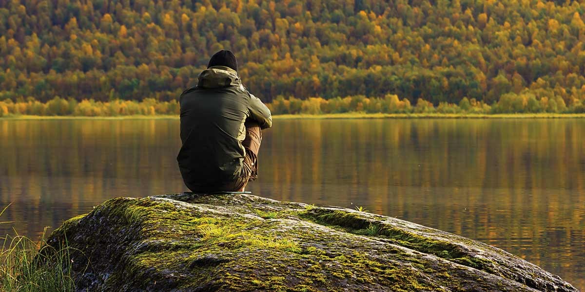 Person sitting on a mossy rock by a lake surrounded by forested hills in autumn colors.