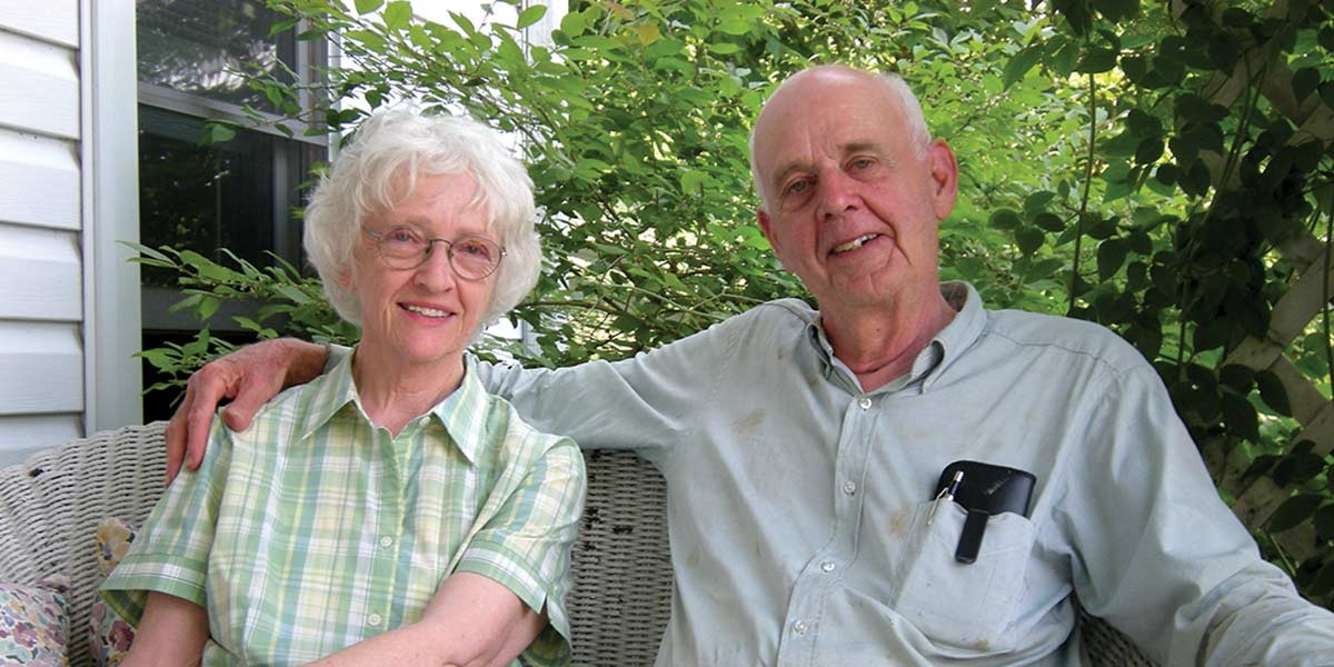 Smiling elderly couple sitting on a white wicker bench surrounded by green foliage.