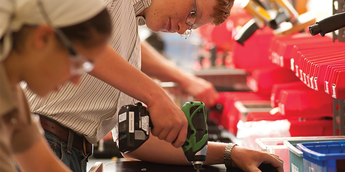 Person using electric drill in workshop with rows of red cases and tools on shelves.