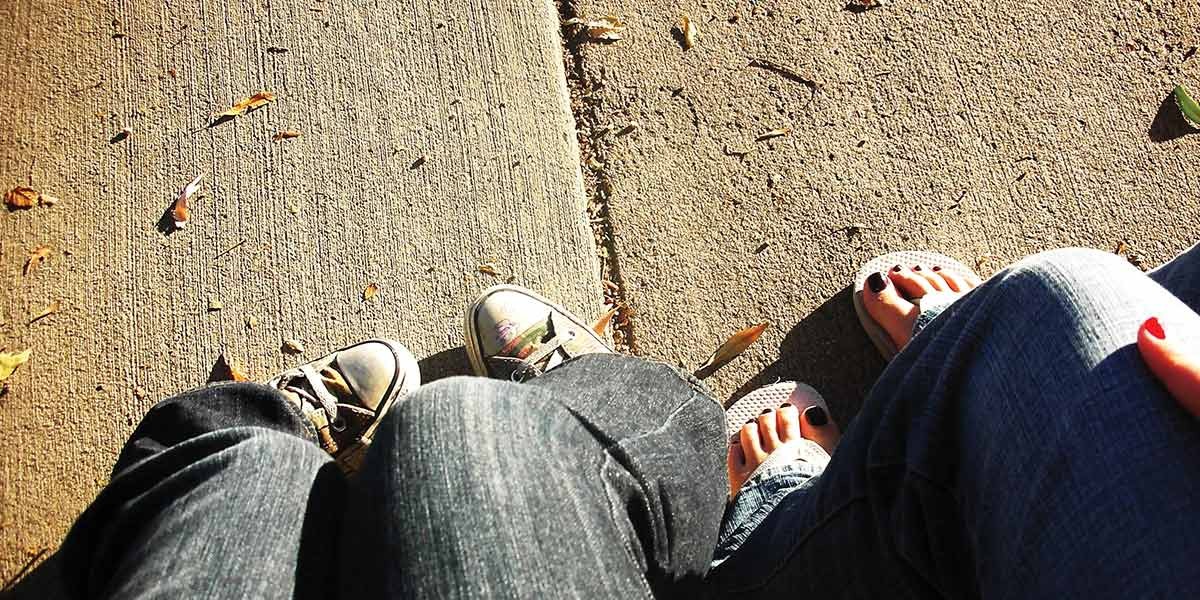 Two people sitting on a sunlit sidewalk, one wearing sneakers and the other flip-flops with painted toenails.