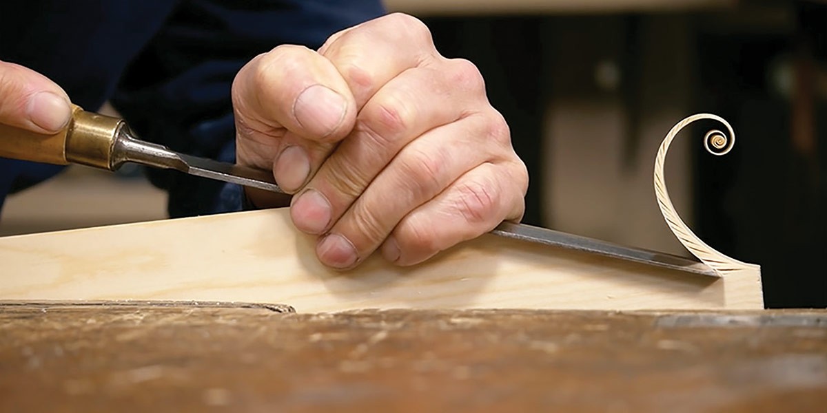 Hand carving a delicate wood curl from a light wood piece on a workbench.
