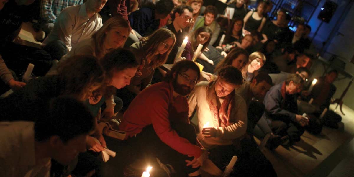 A group of people seated closely indoors, holding lit candles in a dimly lit setting.