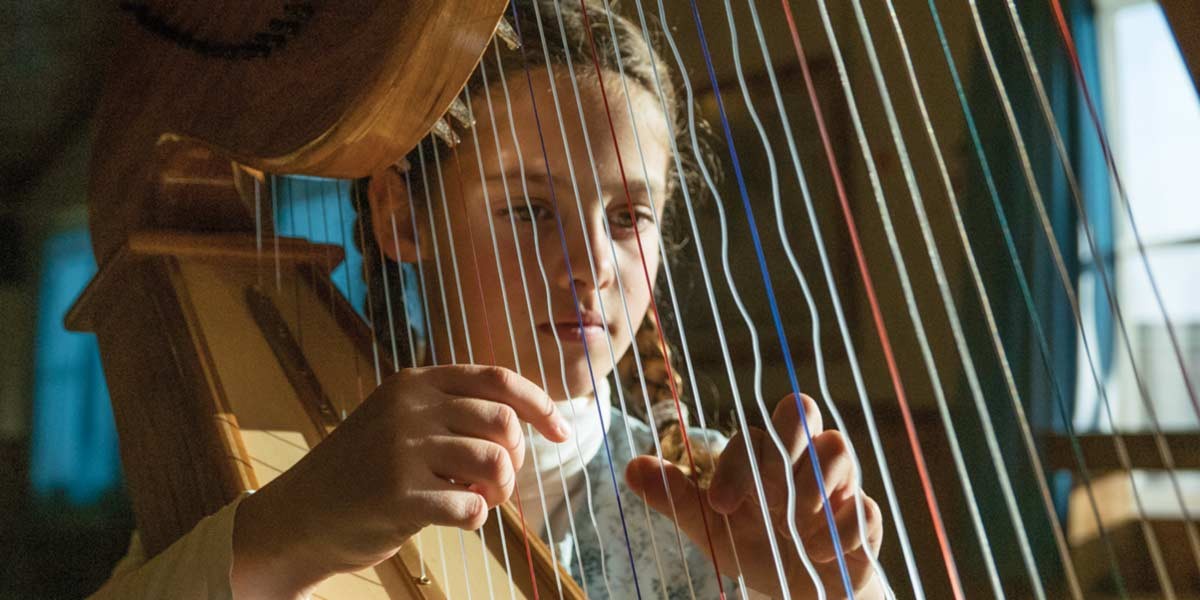 Young girl playing a large wooden harp, focused on plucking the strings indoors near a window.