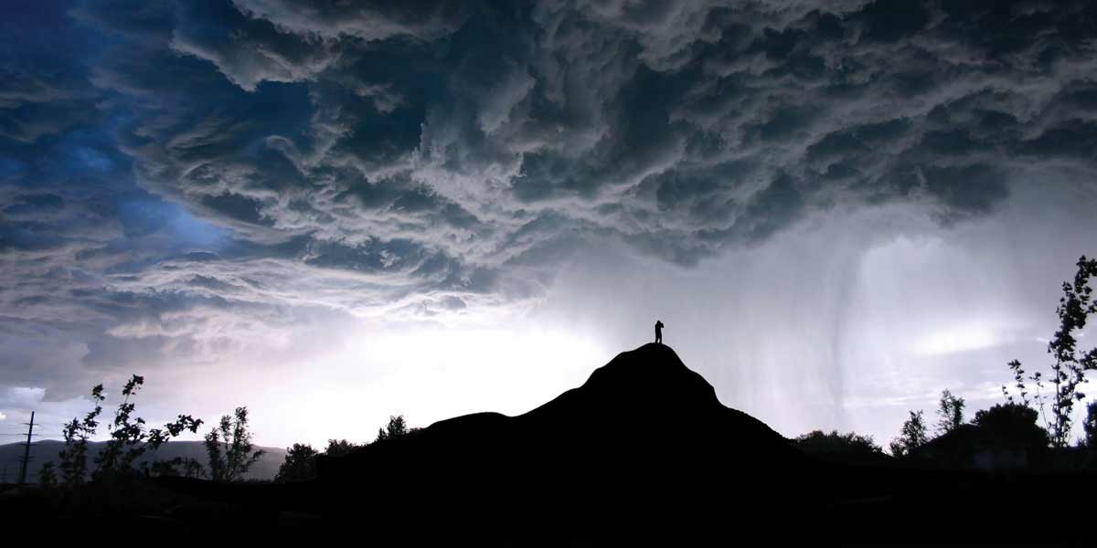 Silhouette of person standing on hilltop under dramatic dark storm clouds at twilight.