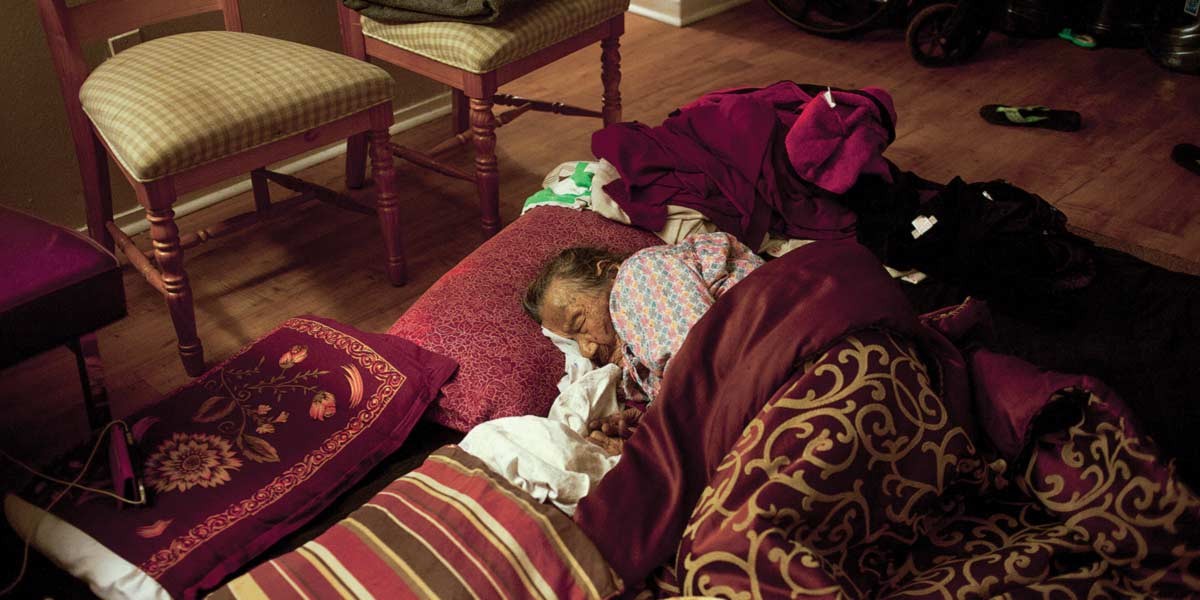 Older person sleeping on the floor, covered with patterned blankets and surrounded by pillows and furniture.