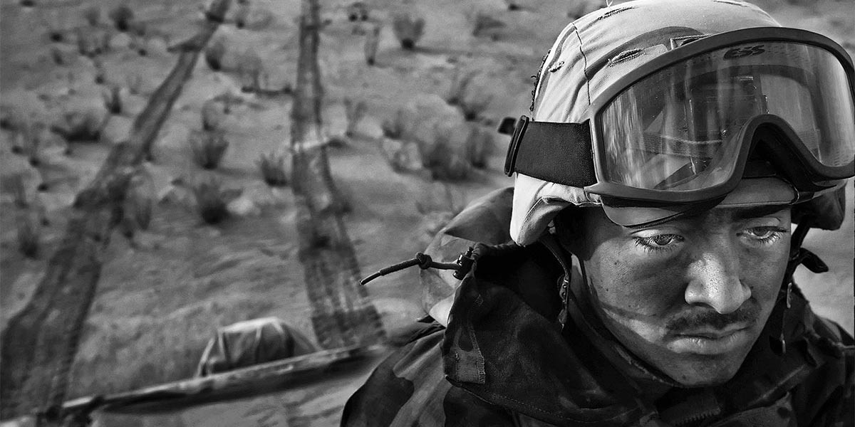 Close-up of soldier in helmet and goggles looking down, with tire tracks and sparse bushes in background.