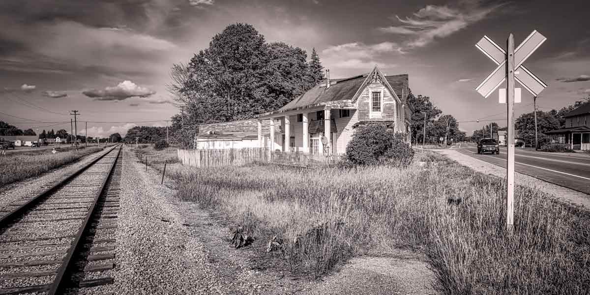 Black and white image of a weathered house near railroad tracks and road with a crossing sign.