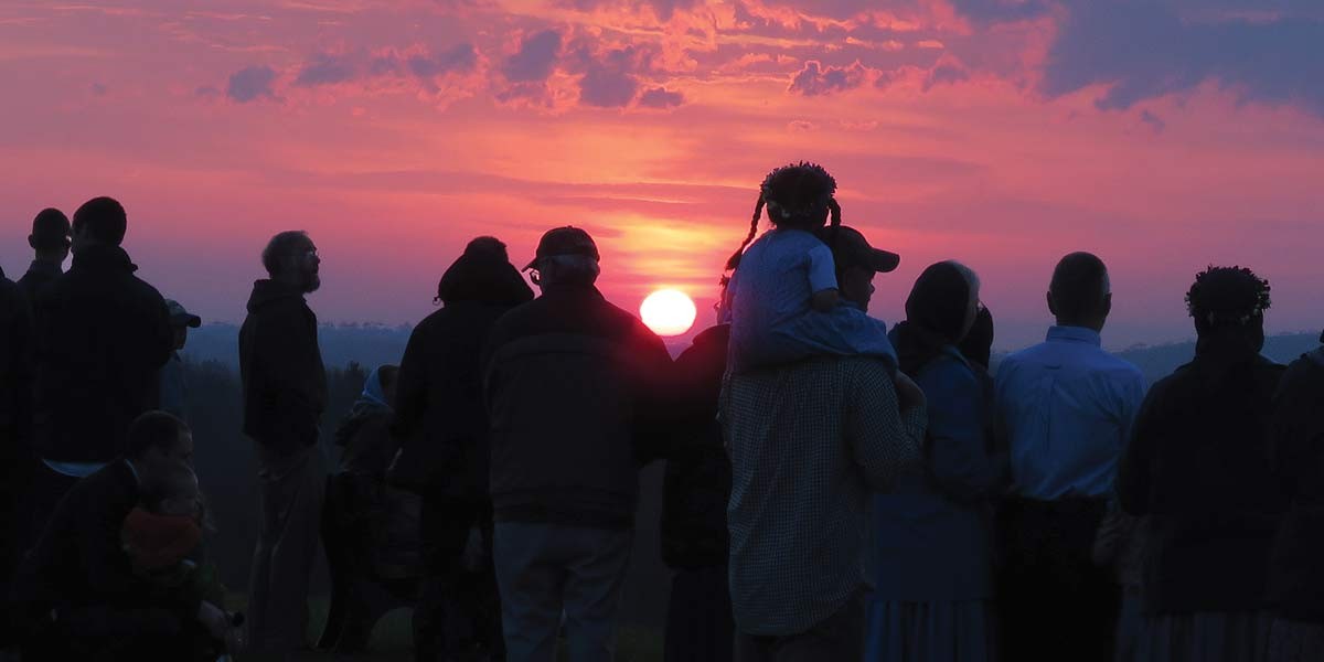 Silhouettes of people gathered outdoors watching a colorful sunset with pink and purple clouds.