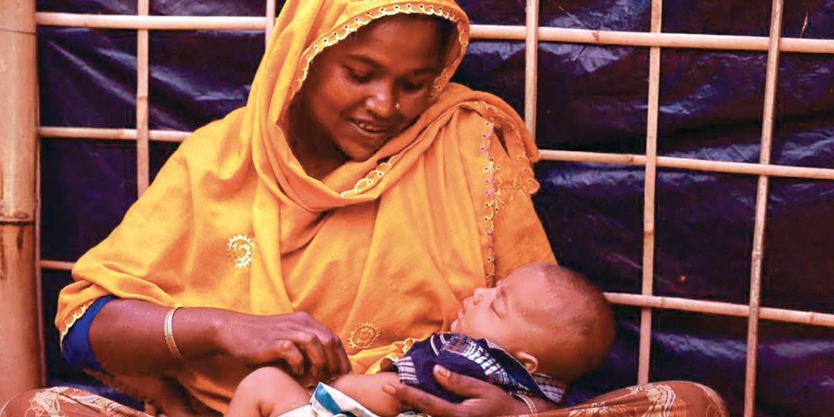 A woman in an orange headscarf cradles and looks lovingly at a sleeping baby in her lap.