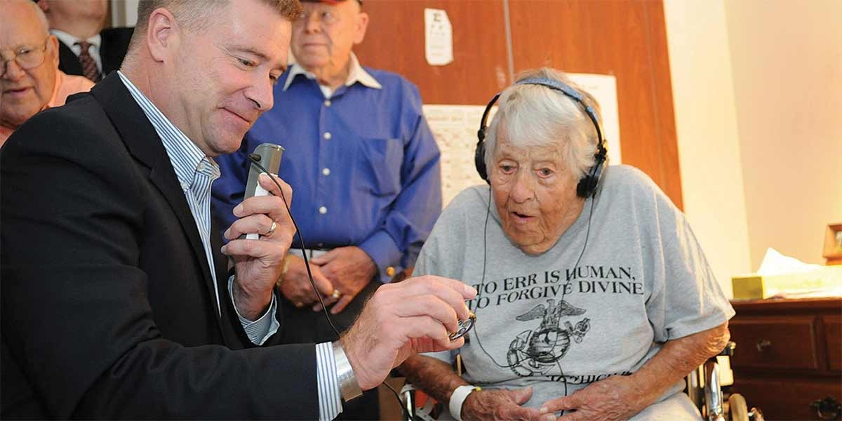 Man demonstrates hearing aid device to elderly woman wearing headphones in a home setting.