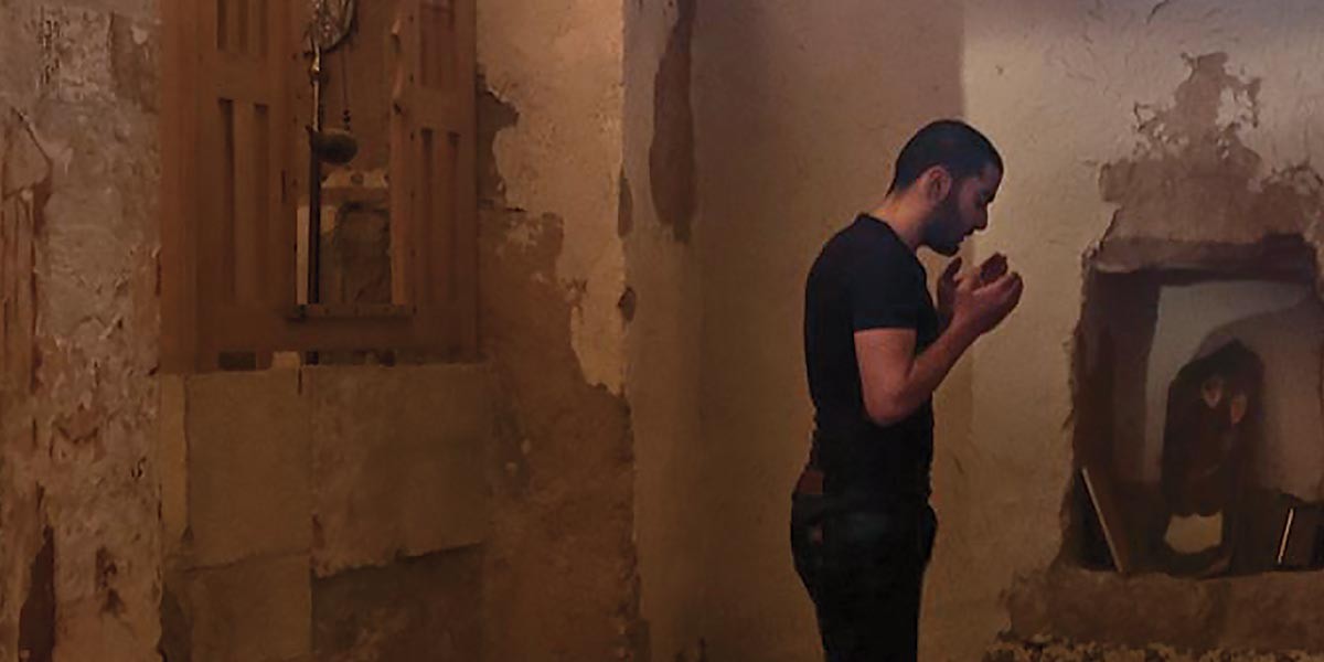 Man in black shirt standing in a rustic room with peeling walls, holding hands up near his face.