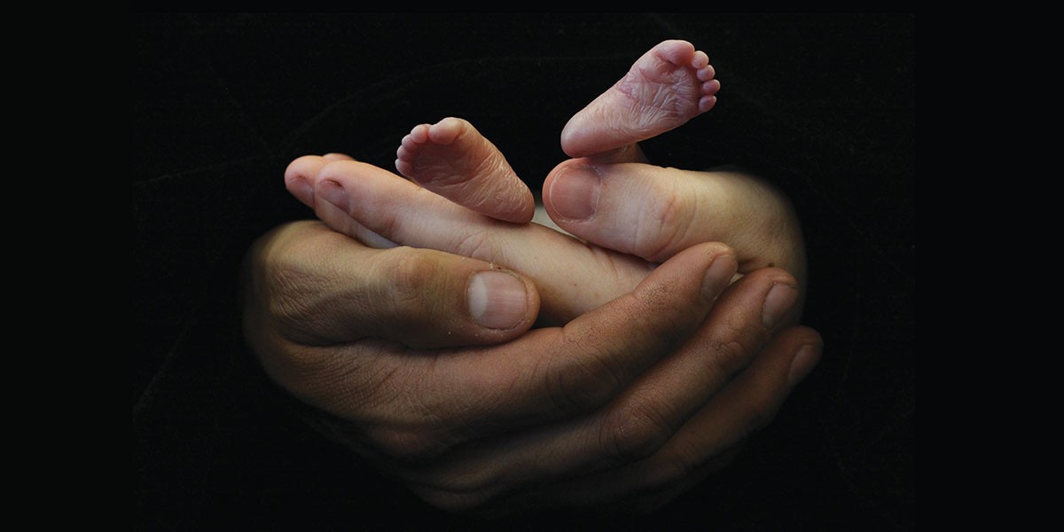 Adult hands gently cradling tiny baby feet against a dark background.