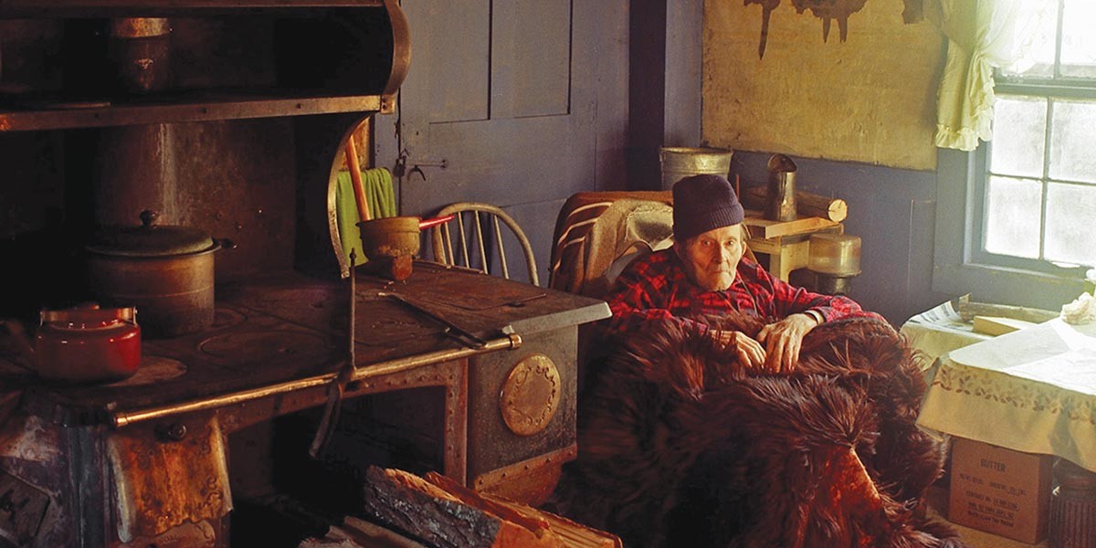 Elderly man in a red plaid shirt and knit cap sitting by a wood stove in a rustic room.