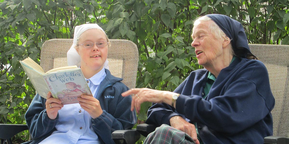 An older woman talks to a young woman reading Charlotte's Web outside with green plants behind them.
