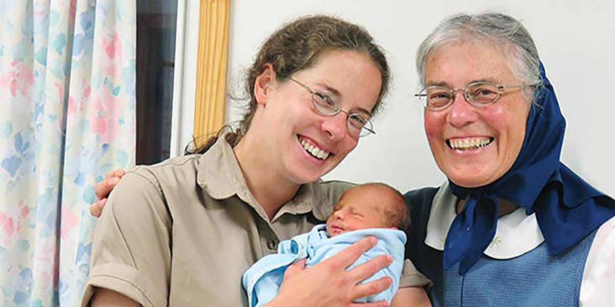 Smiling woman and elderly woman with headscarf holding a newborn baby wrapped in blue cloth.