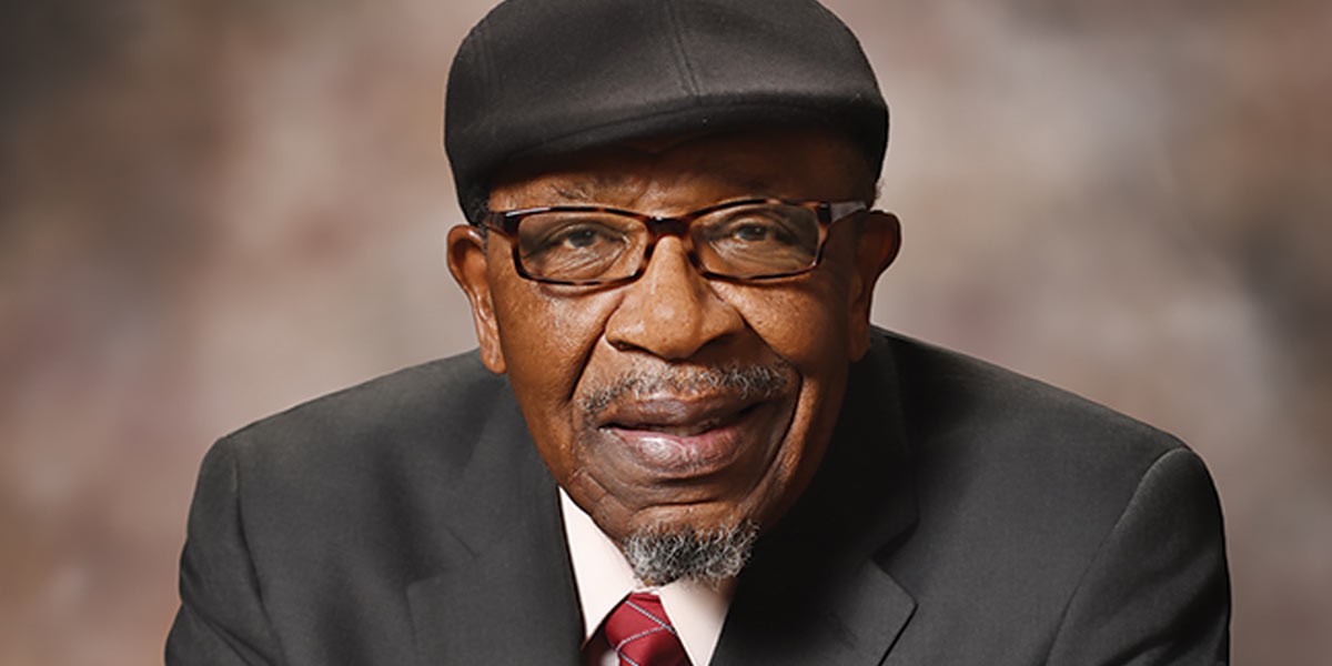 Portrait of an older man wearing glasses, a black cap, suit, and red striped tie smiling gently.