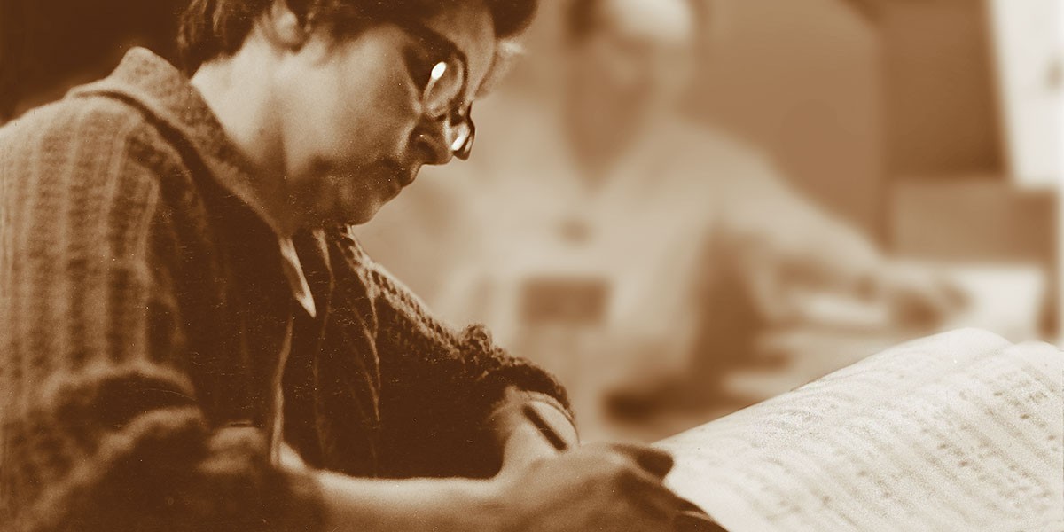Person with glasses closely reading or writing on sheet music with blurred background figure.