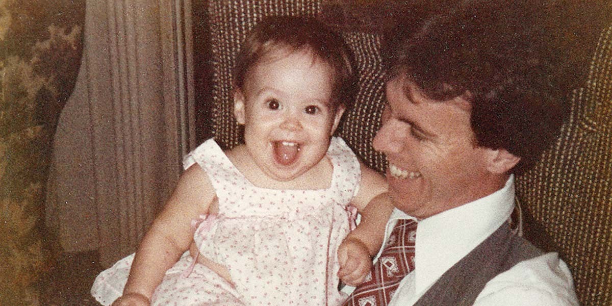 Smiling man in suit holding happy baby girl sticking out her tongue, indoors.