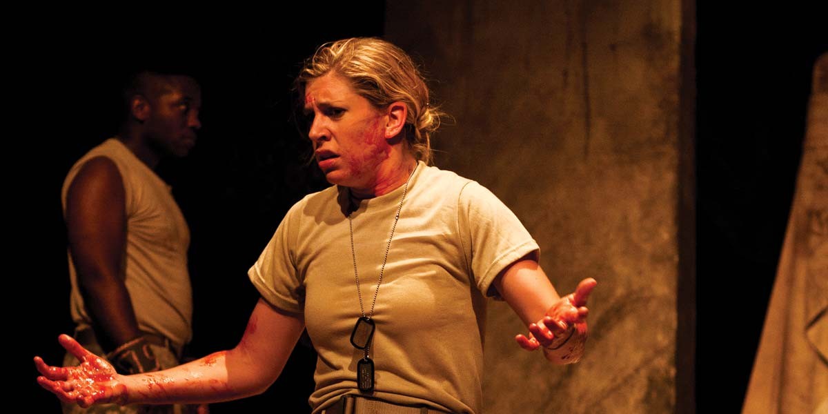Woman with blood on her face and hands wearing dog tags, expressing distress with a man standing blurred in background.