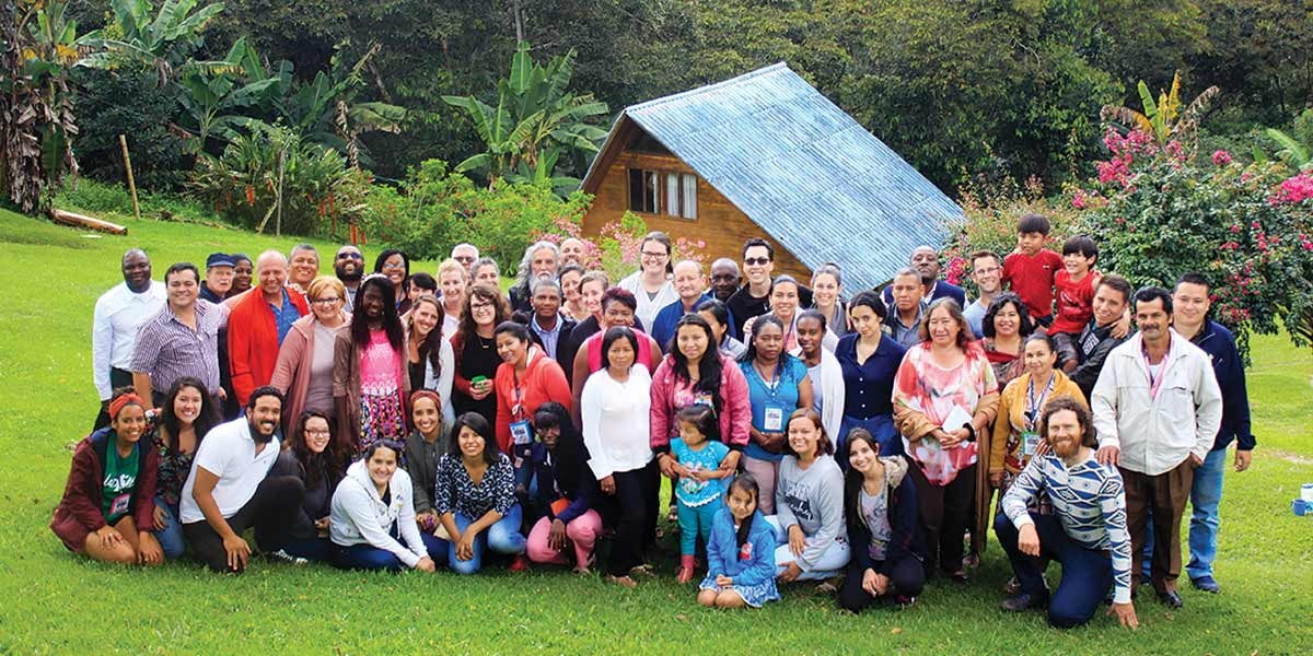 Large diverse group of adults and children posing outside on grass with a small house and trees behind them.