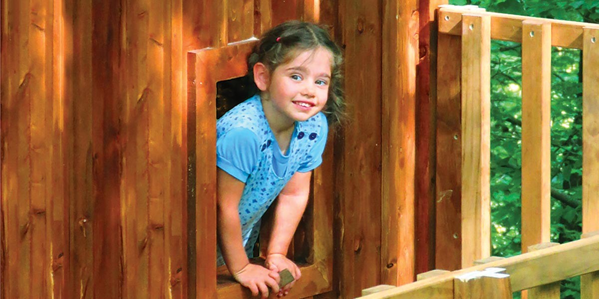 Smiling young girl leaning out of a wooden treehouse window surrounded by green foliage.