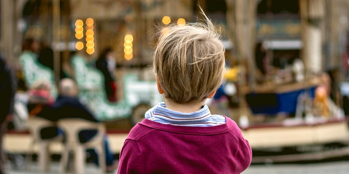 Young boy in a red sweater faces away, looking at blurred lights and carousel in the background.