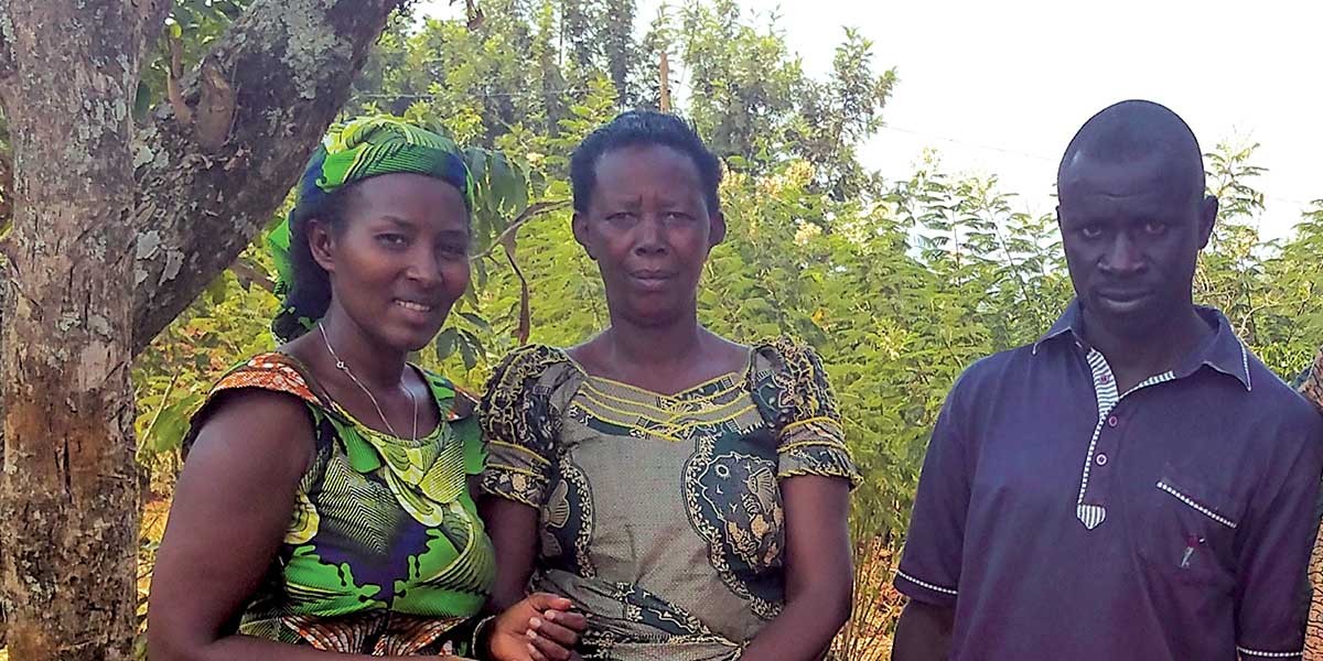 Three people standing outdoors near trees and greenery, two women in patterned outfits and one man in a dark shirt.