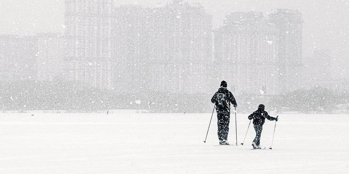 Two people cross-country skiing in heavy snow with city buildings faintly visible in the background.