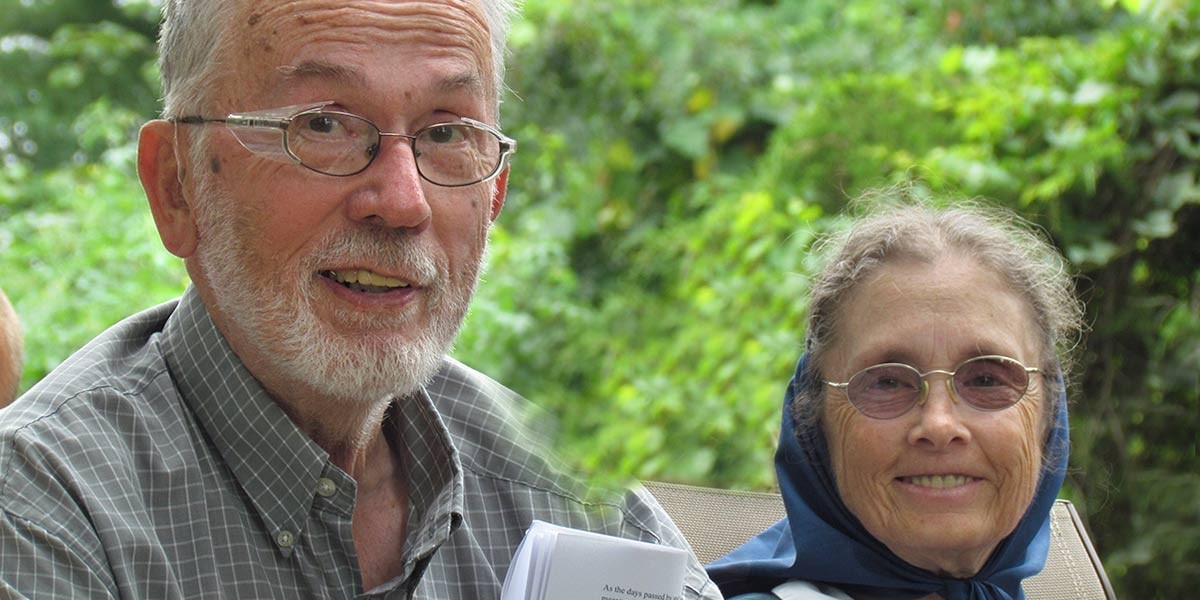 Smiling elderly man with glasses and beard beside elderly woman wearing glasses and a headscarf outdoors.